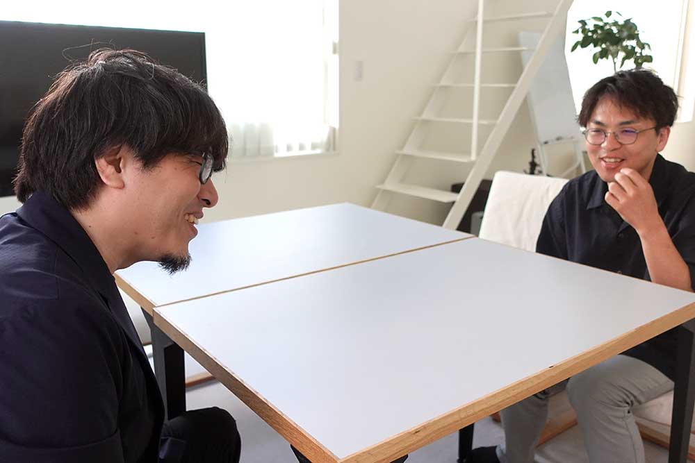 オフィスでテーブルを挟んで話すフジイと倉貫さん。フジイは爆笑、倉貫さんもそれを見て笑顔に