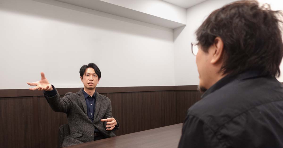 右手を広げて話す神谷さんと、その話を聞くフジイ